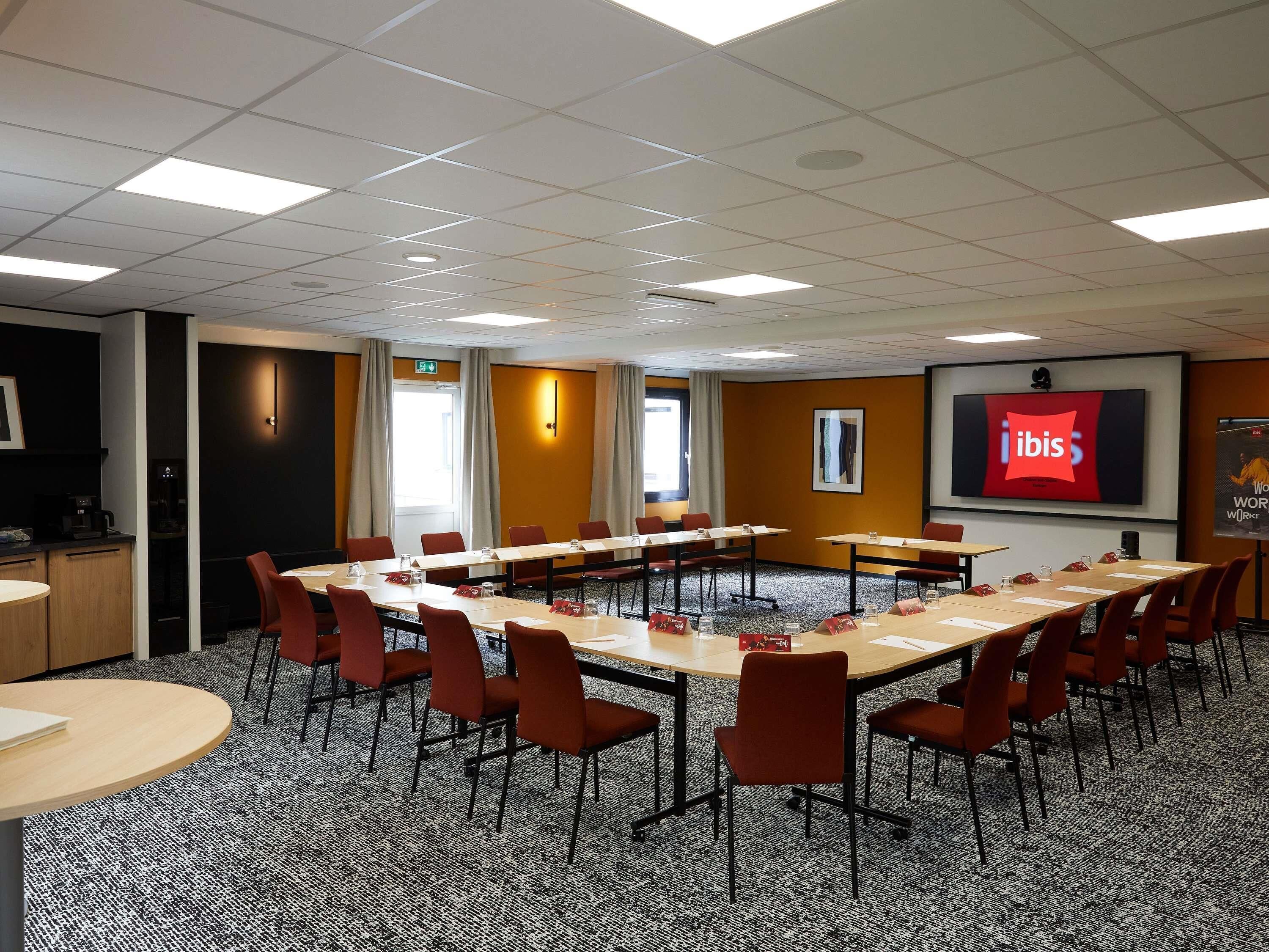 A conference room with a long table, multiple chairs, a projector screen displaying 'ibis', and a coffee machine.