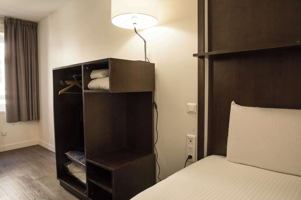 A hotel room with a bed, a wardrobe, and a desk with a lamp.