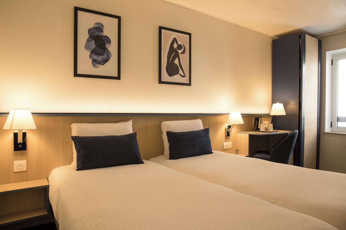 A hotel room with two beds, each with a pillow and a bedside table with a lamp. There are framed artworks on the wall and a window on the right side.