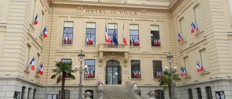 A historic building with a clock tower, named Hotel de Ville, featuring a grand entrance with steps, multiple flags, and classical architecture.