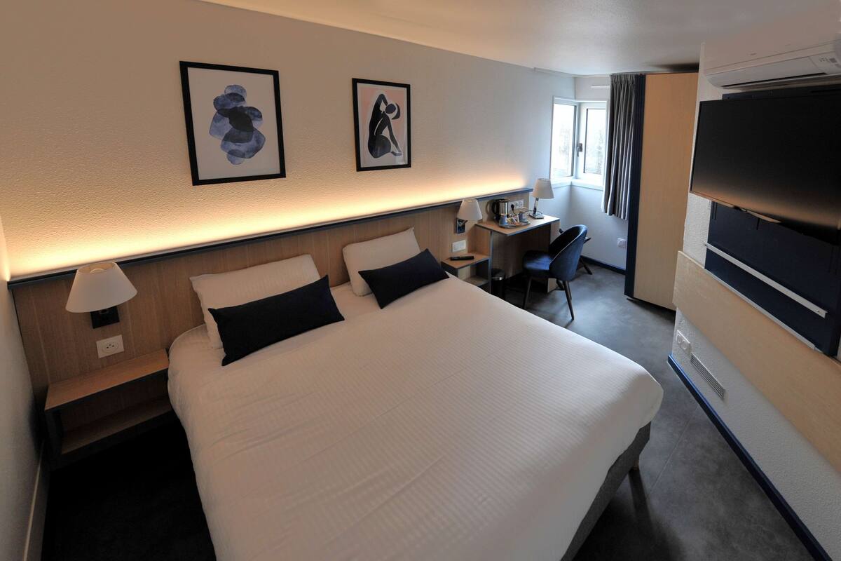 A hotel room with a large bed, bedside tables with lamps, a desk with a chair, a flat-screen TV, and artwork on the wall.