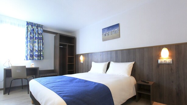 A hotel room with a large bed, a desk with a chair, a wardrobe, and a wall-mounted light fixture. - Brit Hotel Calais (Calais)