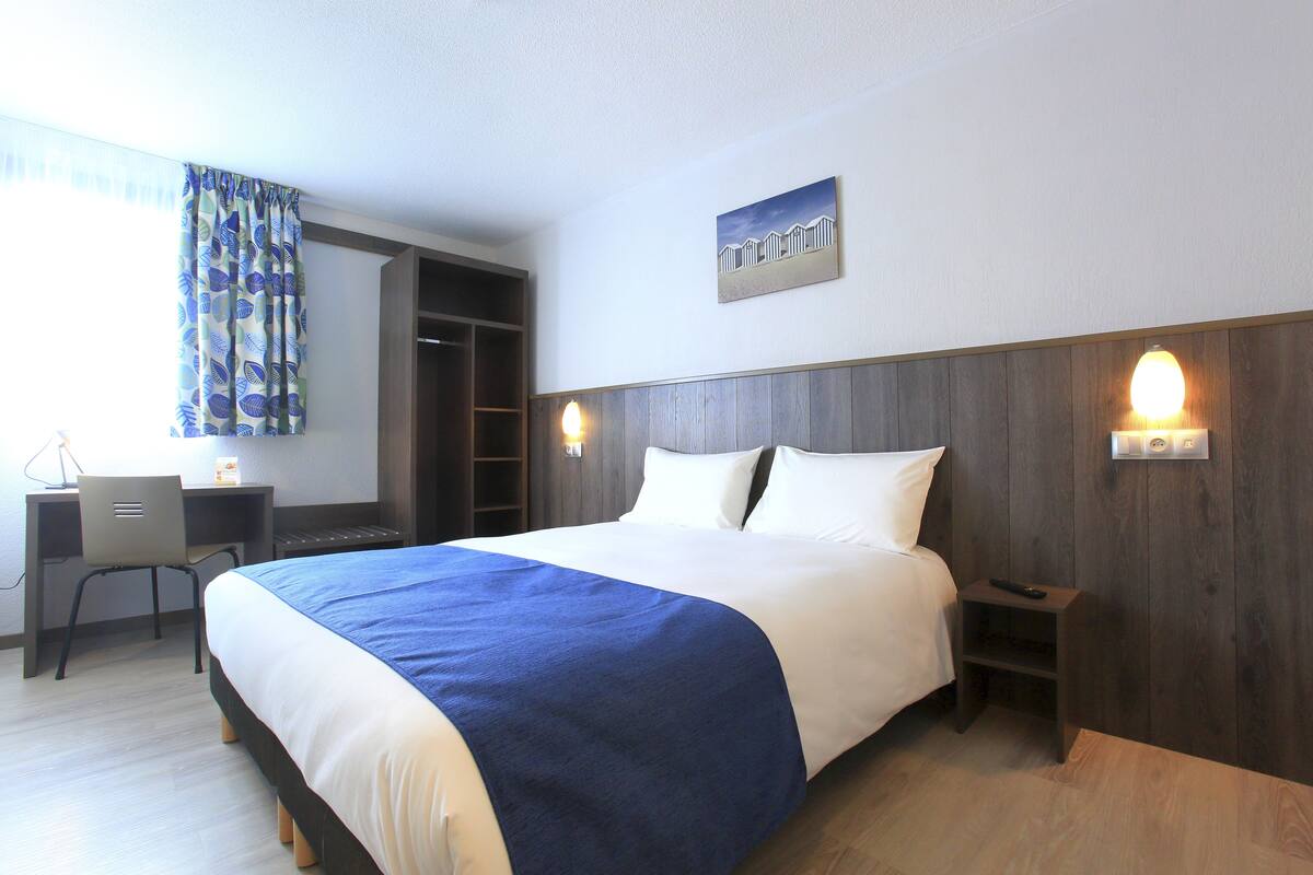 A hotel room with a large bed, a desk with a chair, a wardrobe, and a wall-mounted light fixture.