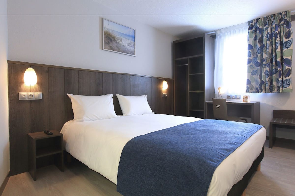 A hotel room with a bed, bedside tables, a desk, a chair, a wardrobe, and a window with curtains.