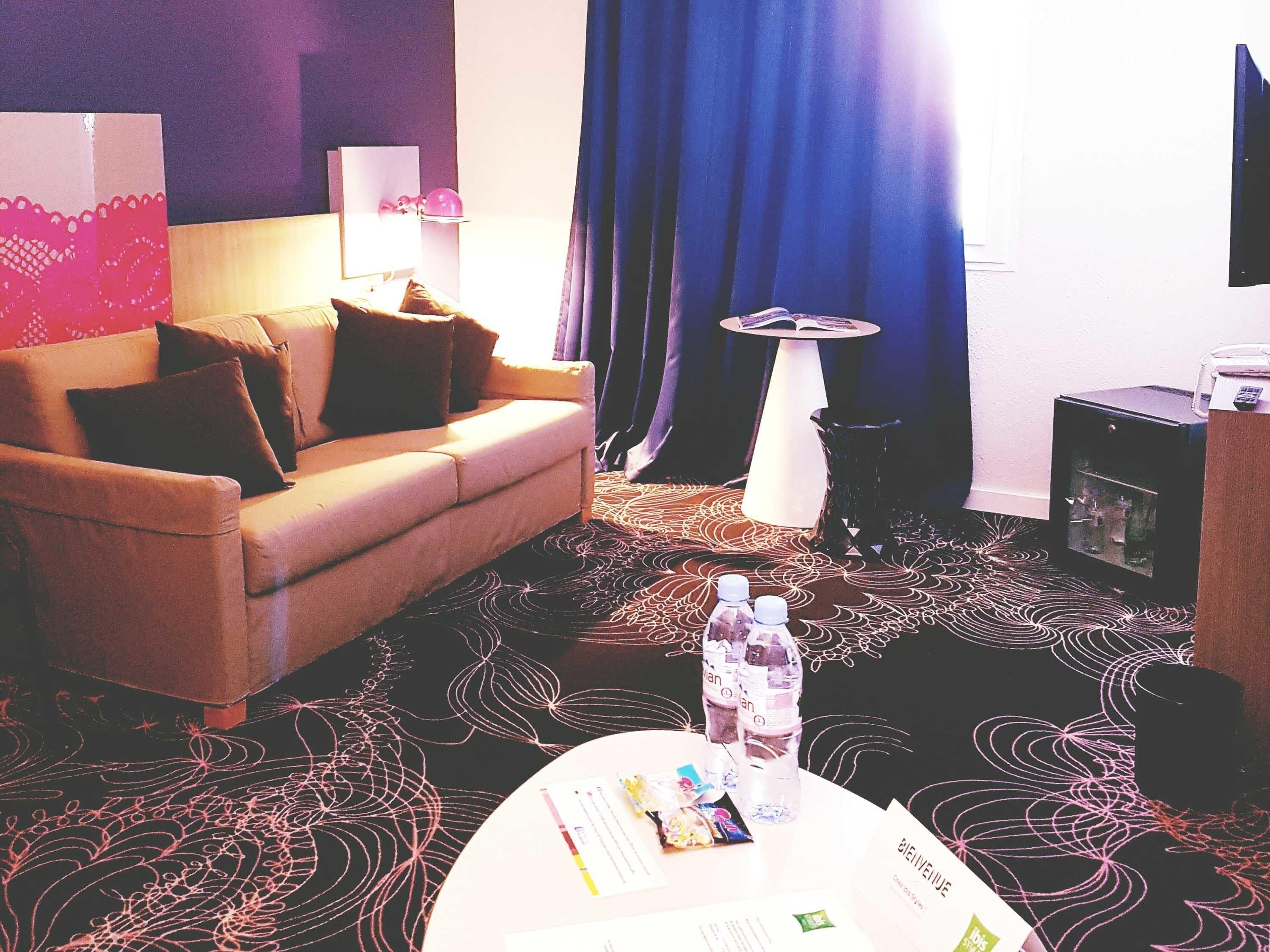 A hotel room with a sofa, a coffee table with items, a small table with a book, and a TV on the wall.