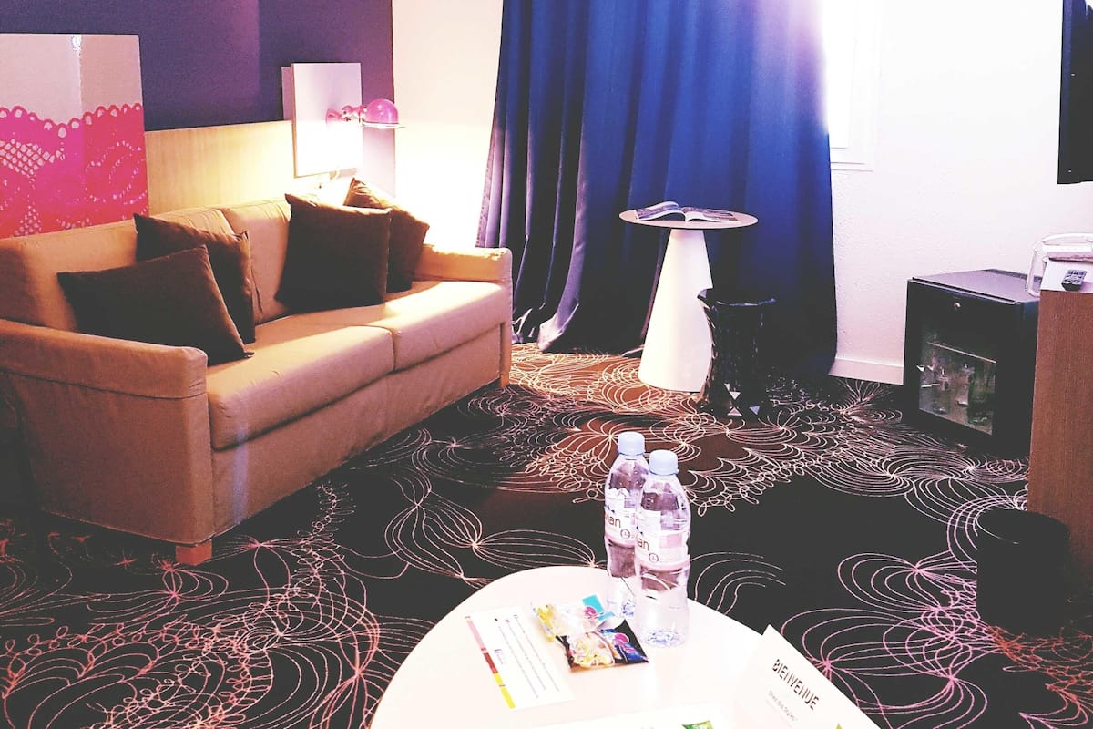 Habitación de hotel con sofá, una mesita de centro con objetos, una pequeña mesa con un libro y un televisor en la pared.