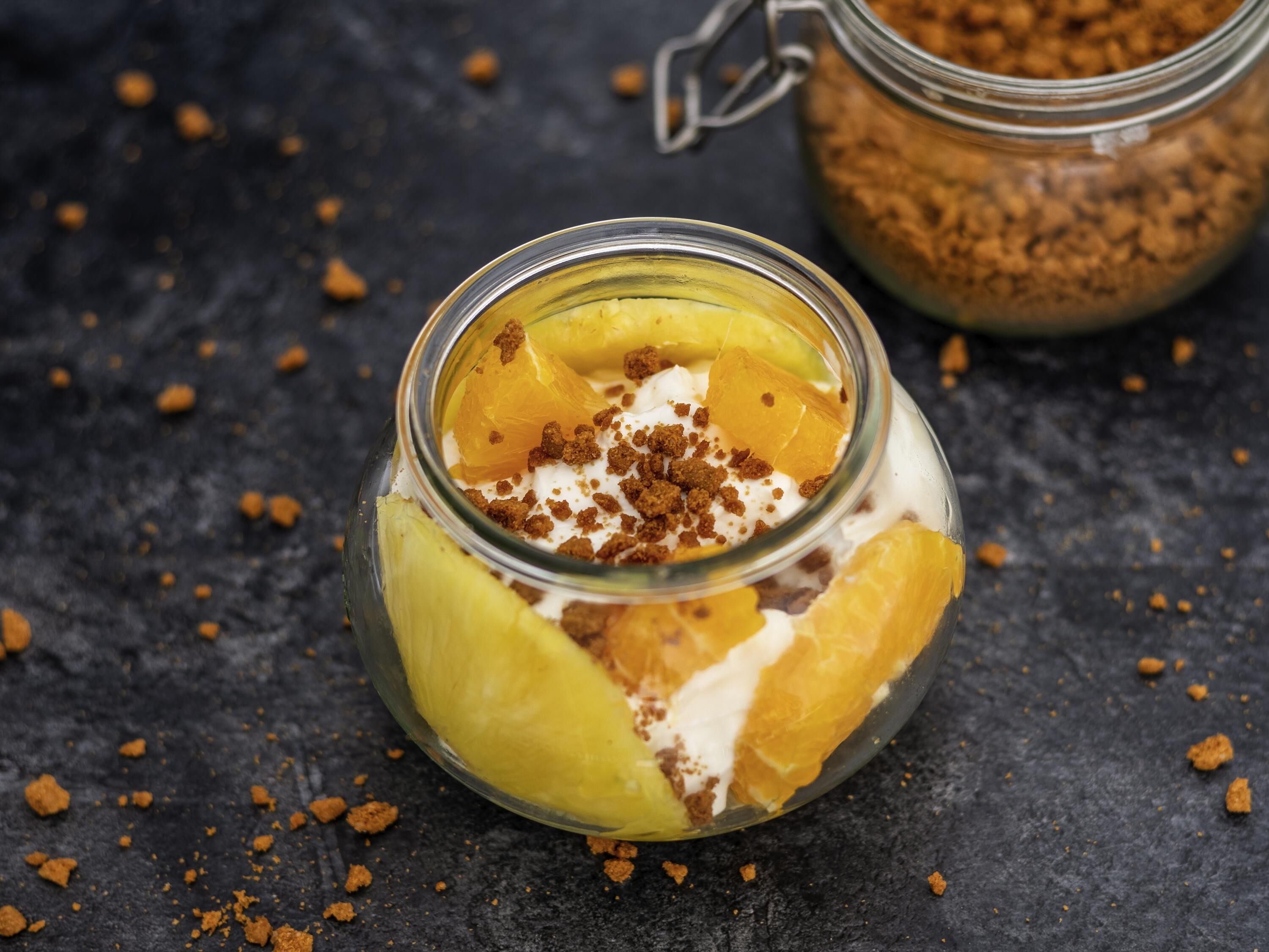A glass jar with a dessert containing fruit and a crumble topping, placed on a dark surface.