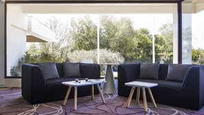 Un área de estar moderna al aire libre con dos sofás negros y dos mesas pequeñas, con vistas a árboles y un edificio al fondo.