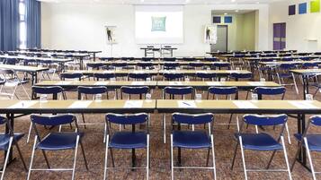 Une salle de conférence avec des rangées de tables et de chaises, un écran de projection et une présentation affichée au mur.