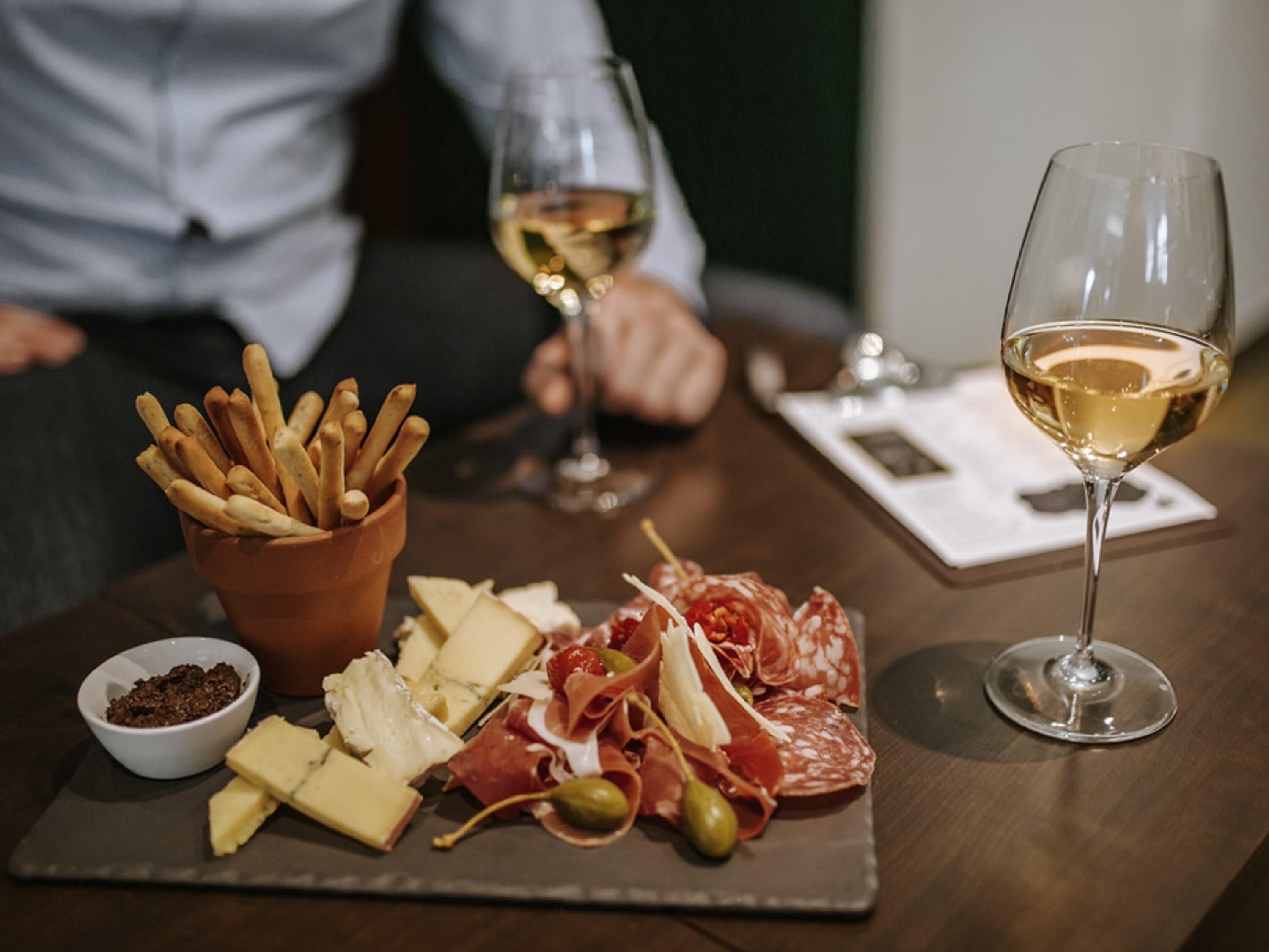 Un tavolo con un tagliere di salumi e formaggi, con olive e grissini, accompagnato da bicchieri di vino bianco.
