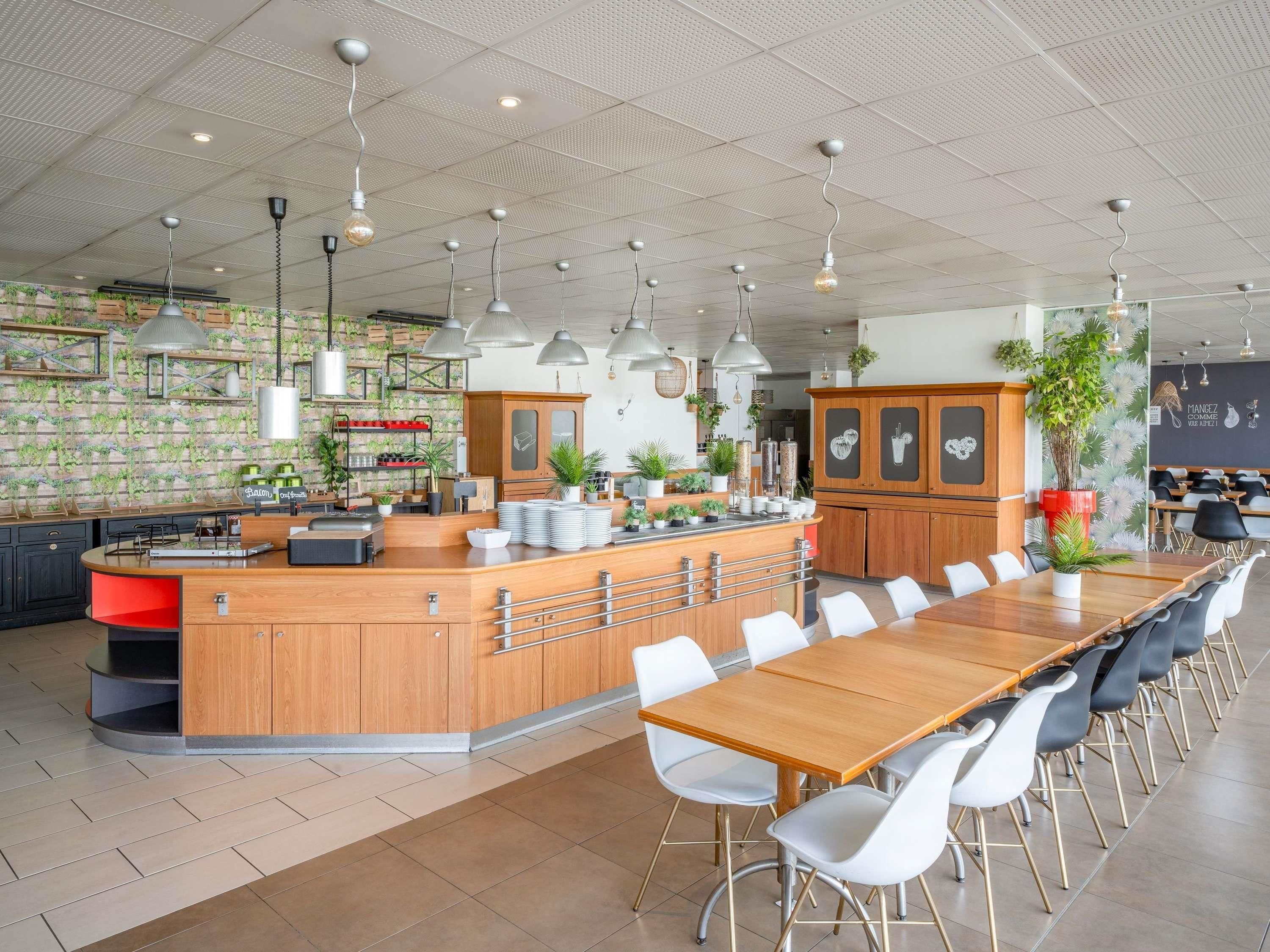 A modern restaurant with a central wooden bar, white chairs, and hanging pendant lights.
