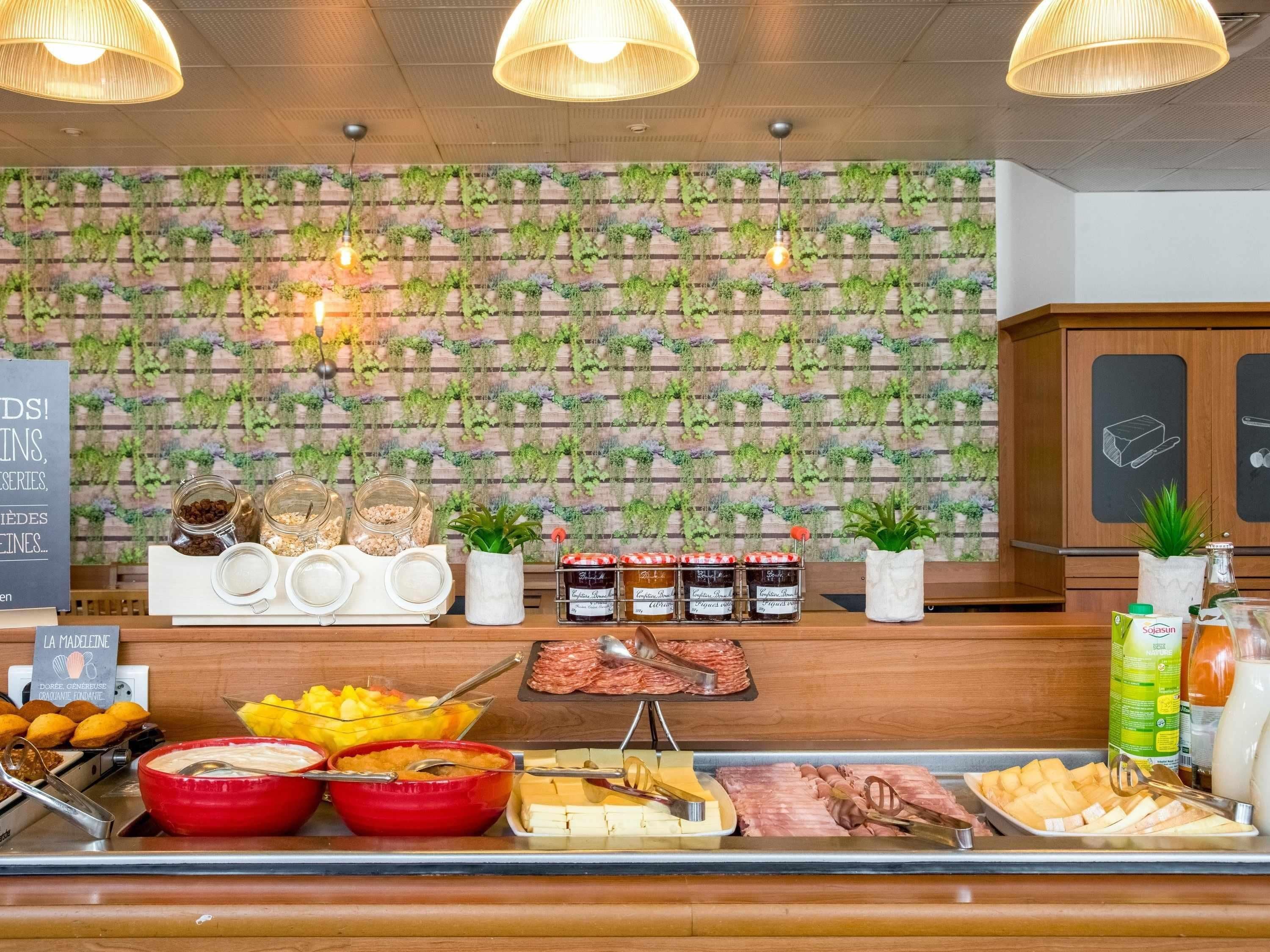 Ett område med en buffé där ett stort urval av matvaror, inklusive frukt, ostar och charkuterier, presenteras på en trädisk med en grön vägg i bakgrunden, där växter hänger ner.