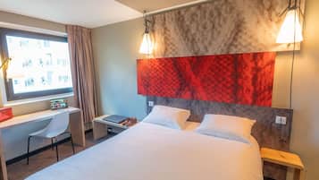 A hotel room with a bed, a desk with a laptop, a chair, and a large window.
