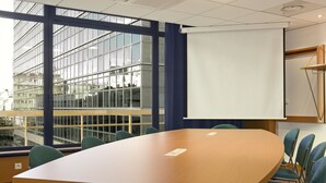 Une salle de conférence équipée d’une grande table, de chaises, d’un écran de projection et de grandes fenêtres donnant sur un immeuble.