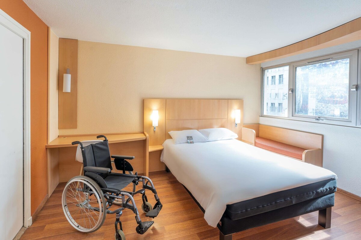 Hotelkamer met een rolstoel, een bed, nachtkastje en een raam met uitzicht.
