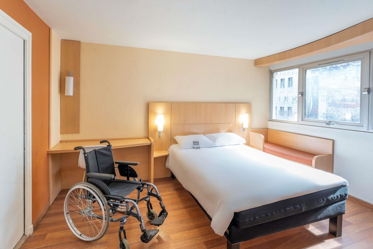 Une chambre d’hôtel équipée d’un fauteuil roulant, d’un lit, d’une table de chevet et d’une commode, ainsi que d’une fenêtre donnant sur un paysage agréable.