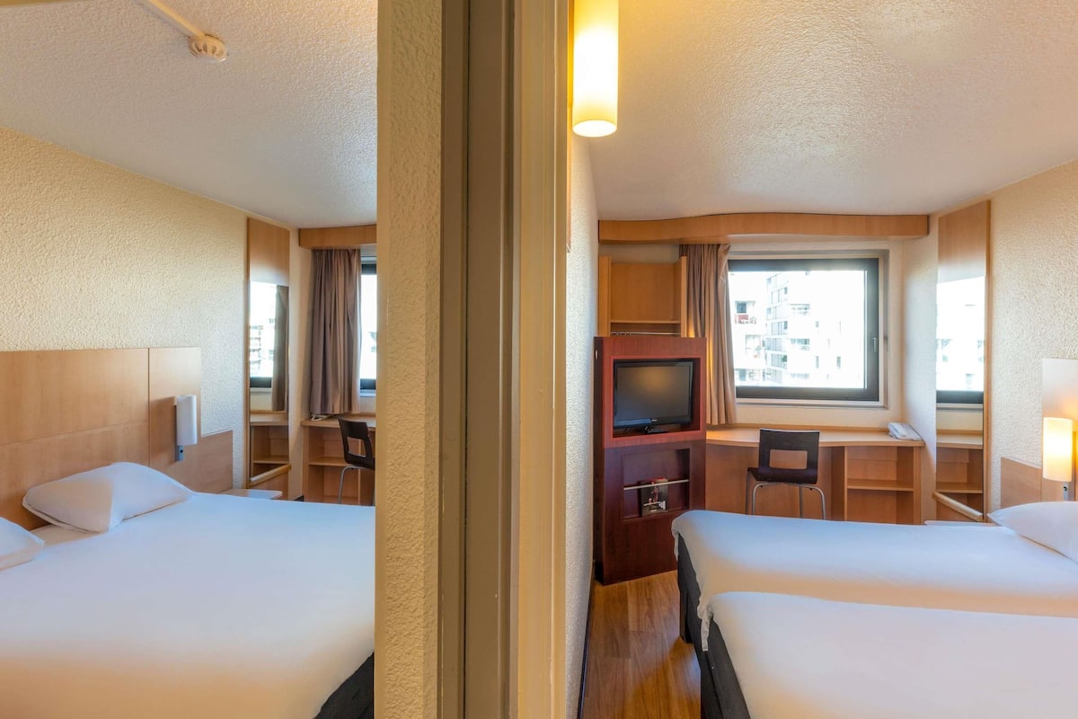 Une chambre d’hôtel avec deux lits, une télévision, un bureau et une fenêtre donnant sur des immeubles.