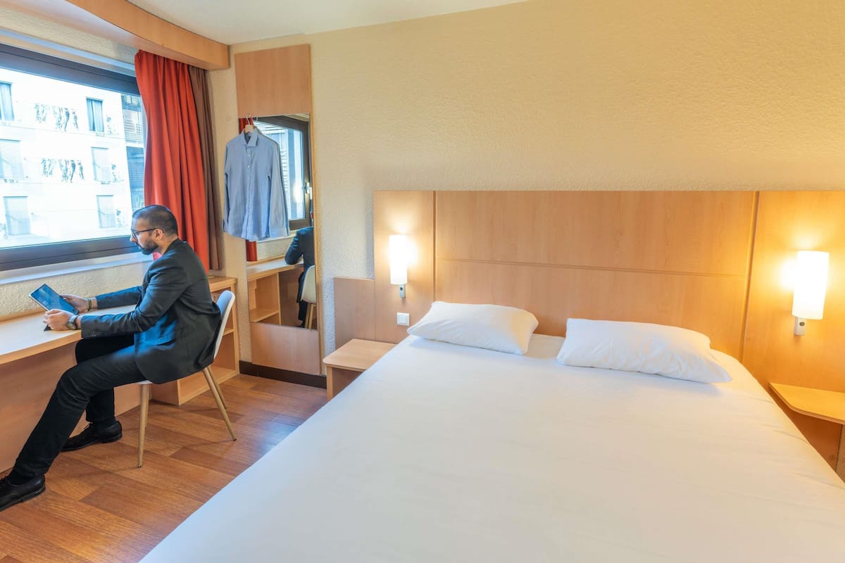 Une chambre d’hôtel avec un lit, un bureau, une chaise et une fenêtre avec des rideaux.