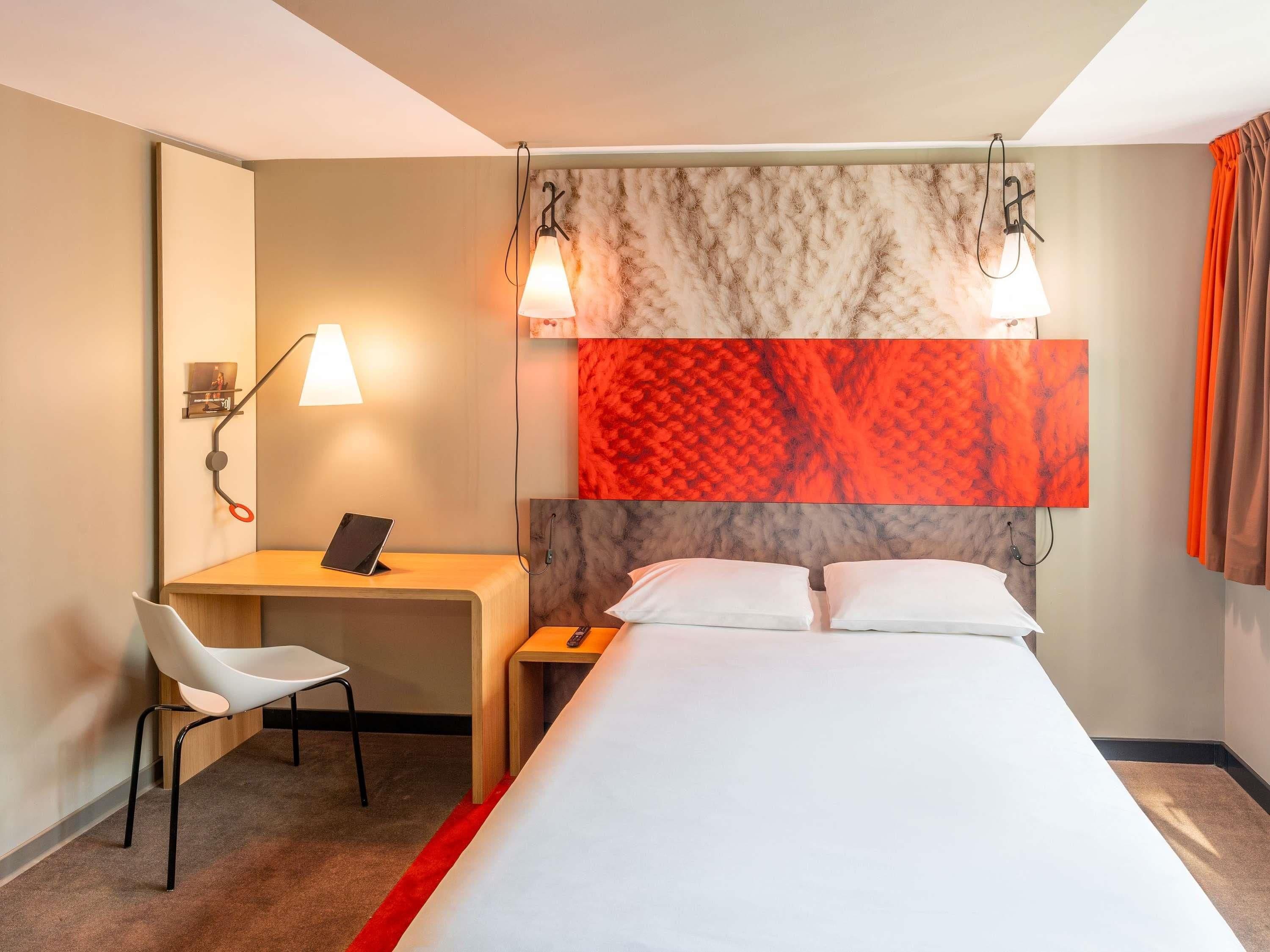 A hotel room with a bed, a desk with a chair, a lamp, and a red carpet.