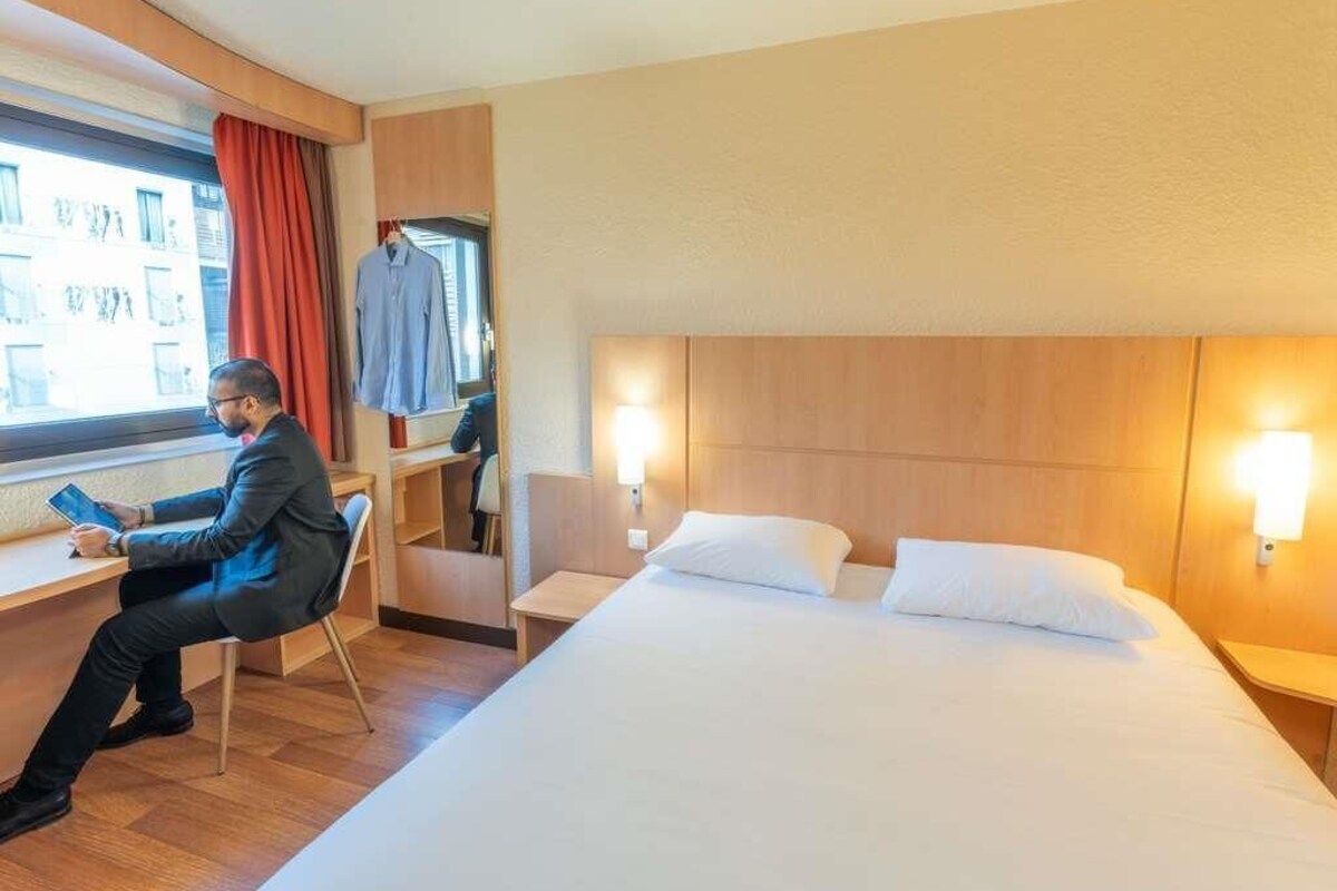 A hotel room with a bed, a desk, a chair, a window with curtains, and a wall-mounted light.