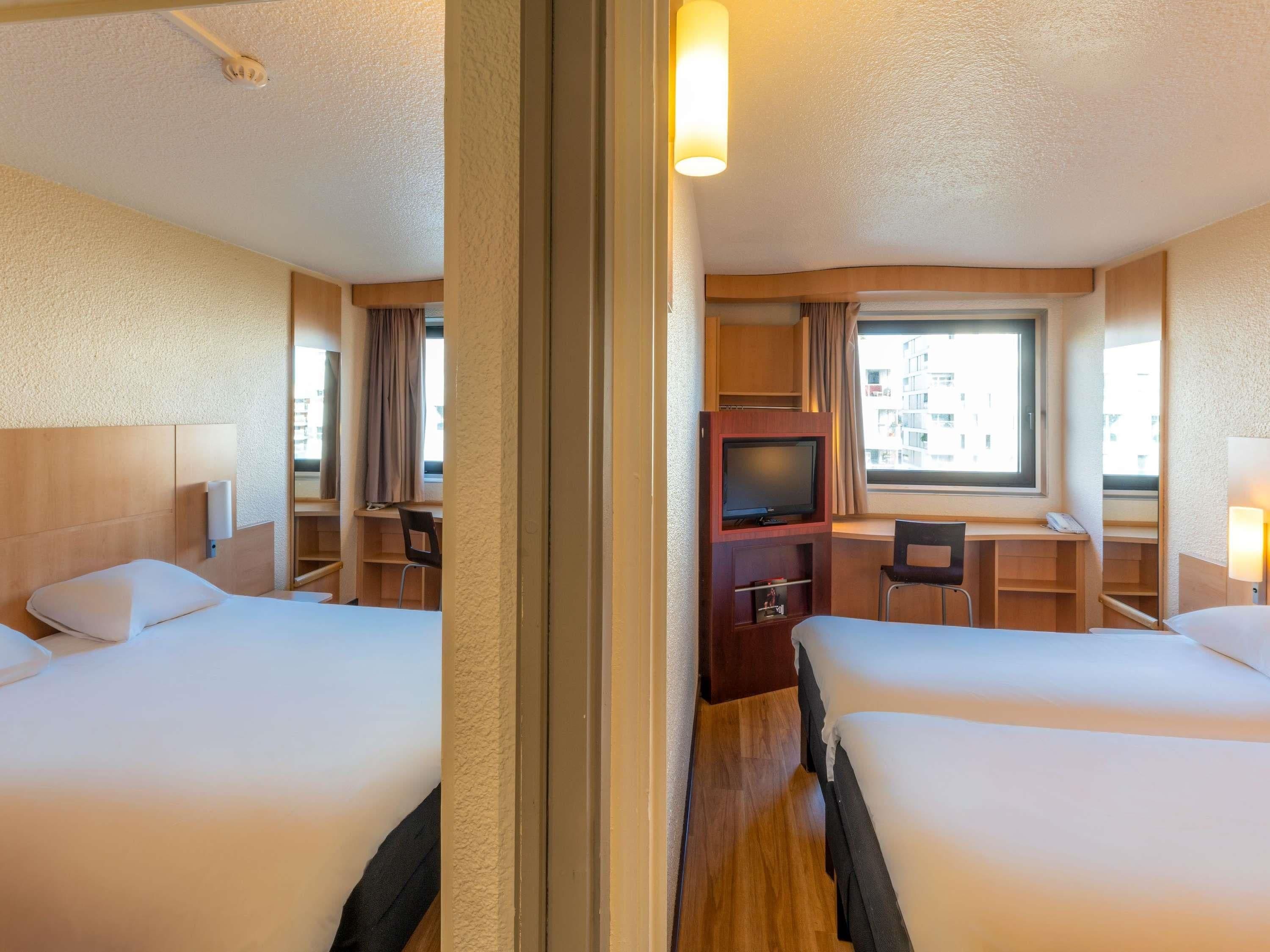 A hotel room with two beds, a TV, a desk, and a window with a view of buildings.