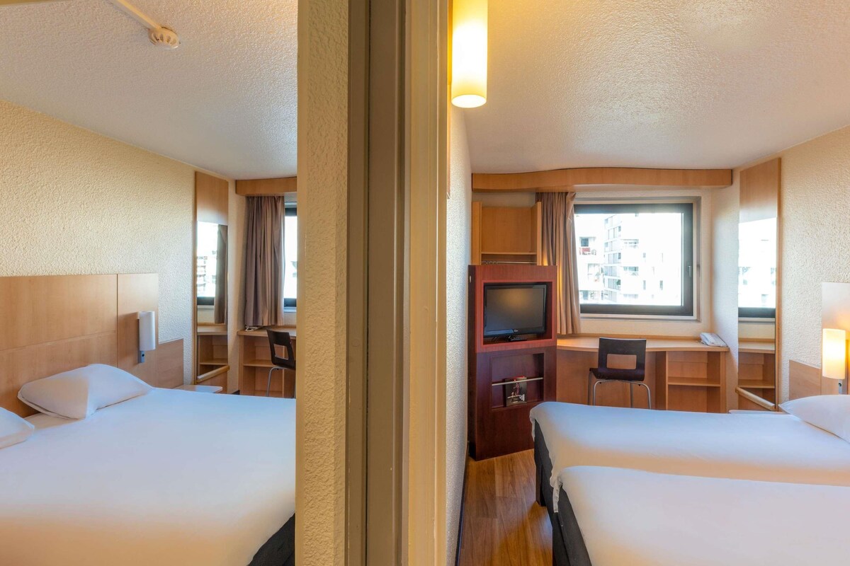Een hotelkamer met twee bedden, een televisie, een bureau en een raam met uitzicht op gebouwen.