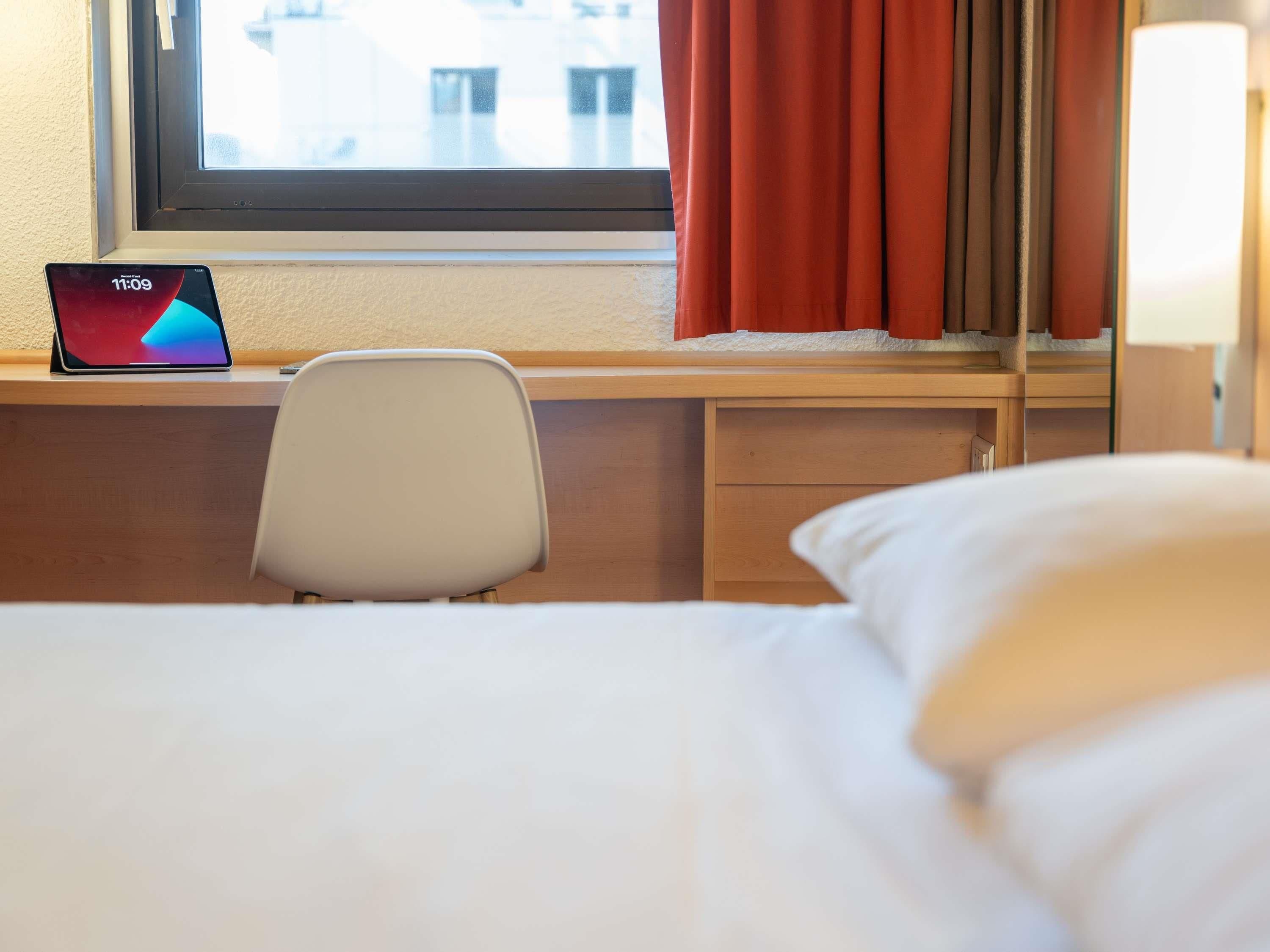 Quarto de hotel com uma cama, uma mesa com um laptop, uma cadeira e uma janela com cortinas.
