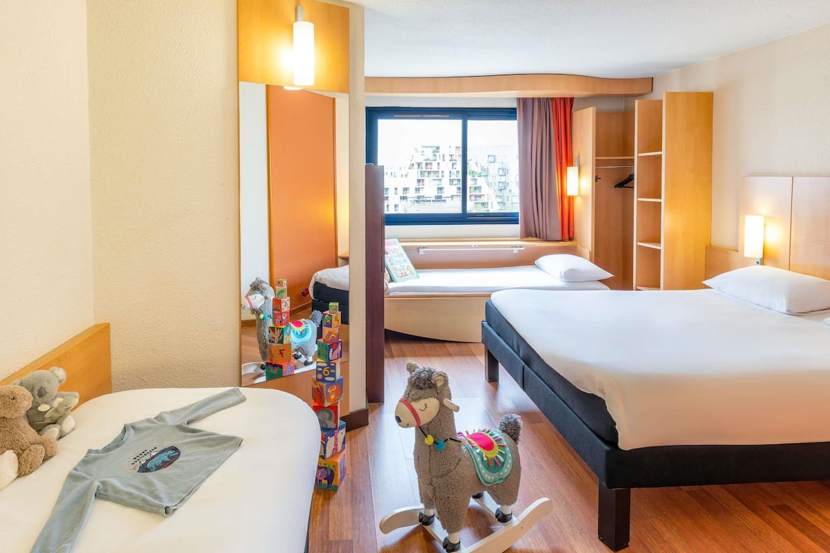 Une chambre d’hôtel avec deux lits, une grande fenêtre donnant sur des immeubles, une armoire en bois et un ours en peluche sur l’un des lits.