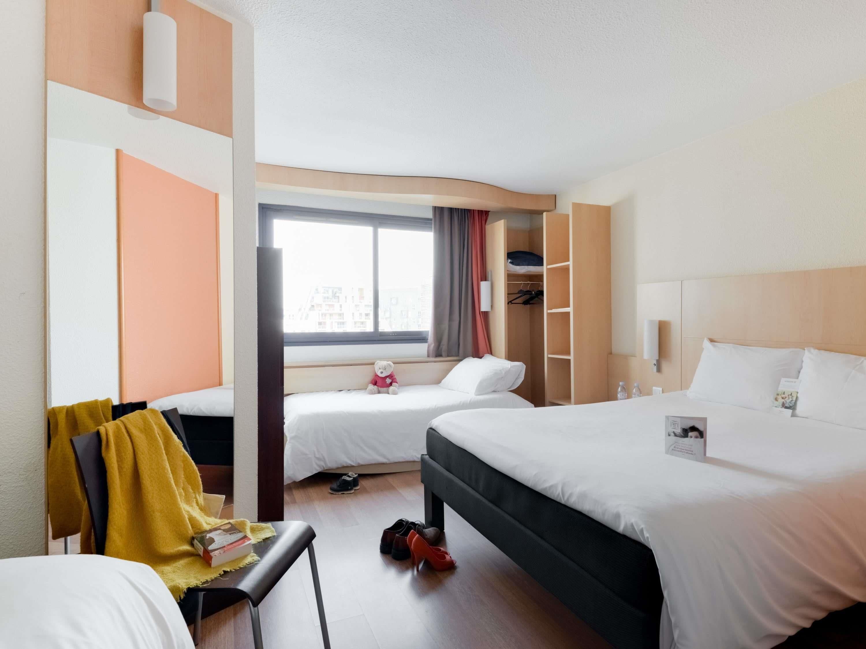 Habitación de hotel con dos camas, una ventana con cortinas, un armario y una silla con una manta amarilla.