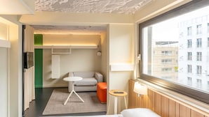 A hotel room with a bed, a small table, a sofa, a window with a view of buildings, and a ceiling with a patterned design. - ibis Paris 17 Clichy-Batignolles (Paris)