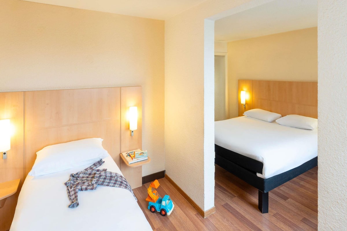 Une chambre d’hôtel avec deux lits, des têtes de lit en bois, des lampes de chevet, une table de chevet avec un livre et un camion jouet sur le sol.