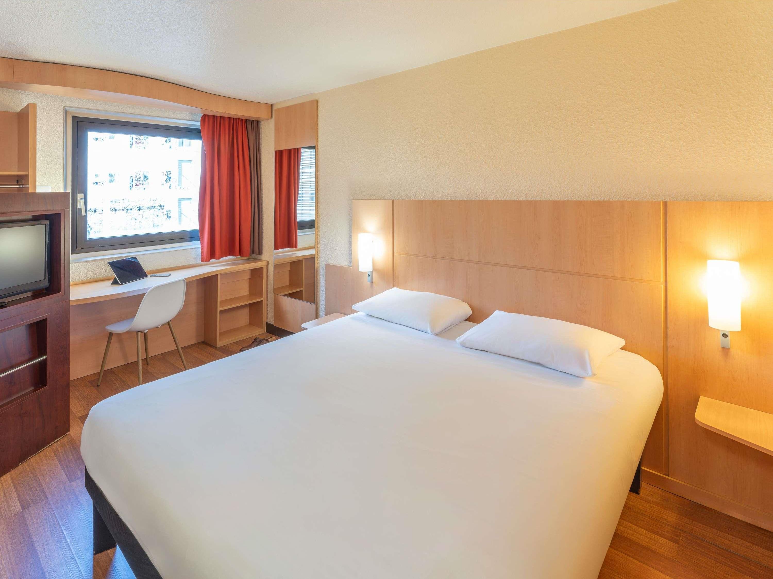 A hotel room with a large bed, a desk with a laptop, a TV, and a window with curtains.