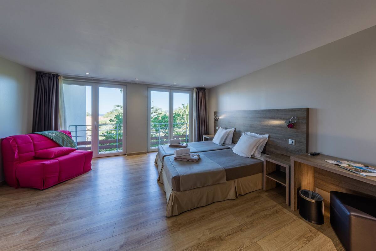 A modern hotel room with two beds, a desk, a chair, and a balcony with a view of greenery.