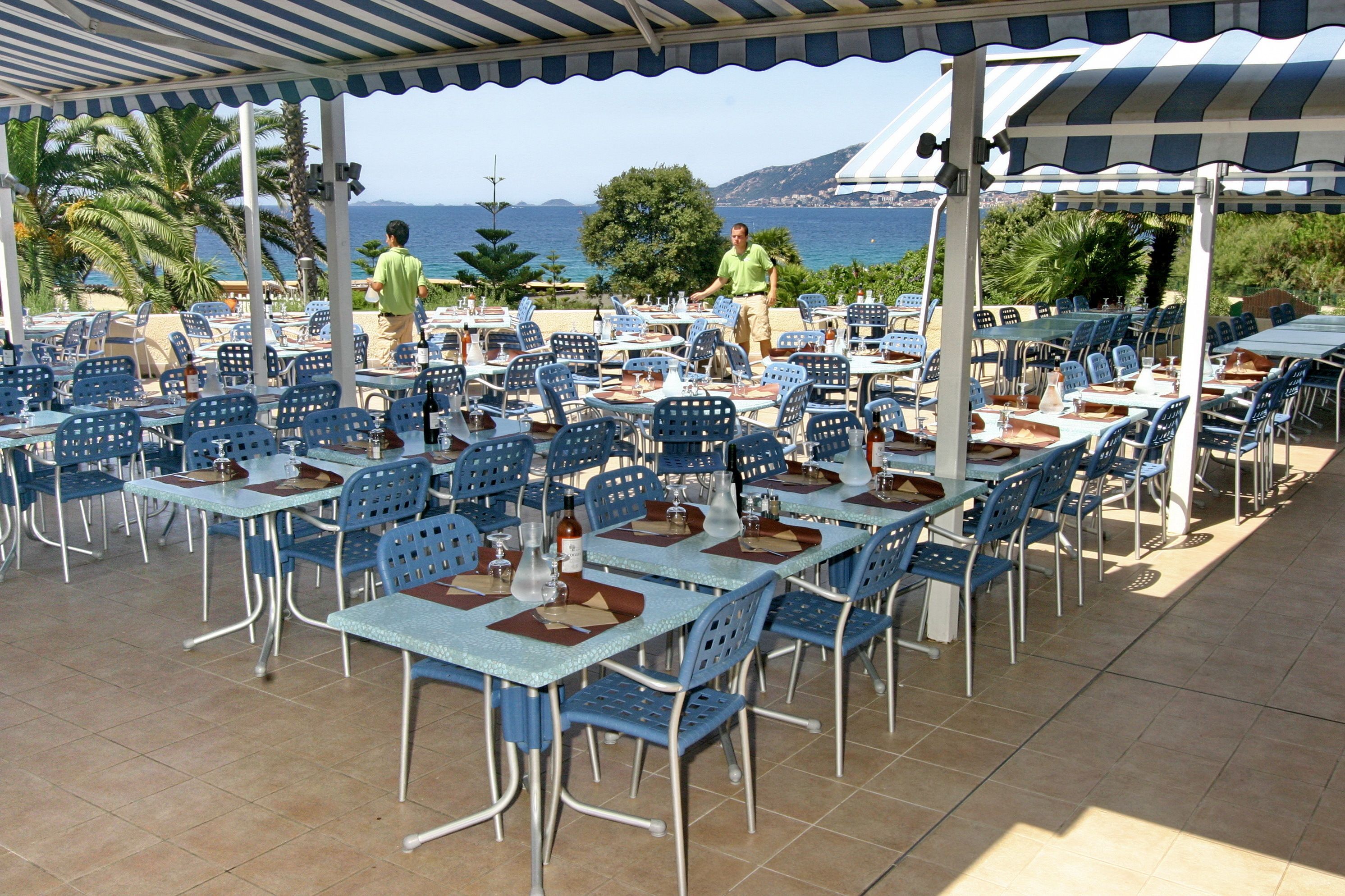 An outdoor dining area with blue chairs and tables, a covered section, and a view of the sea and mountains.