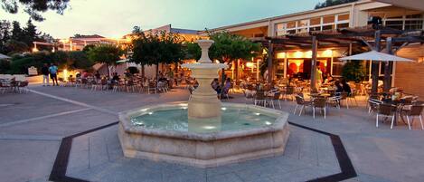 An outdoor cafe area with a central fountain, surrounded by outdoor seating and a building in the background.