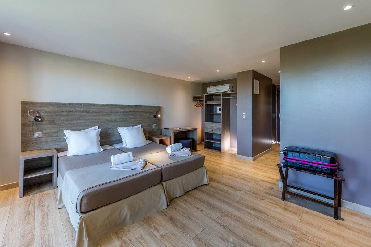 A modern hotel room with a large bed, bedside tables, a wardrobe, and a suitcase on a stand.
