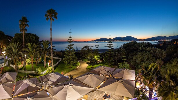 Un resort con sombrillas exteriores, palmeras y vistas al mar y las montañas al atardecer.