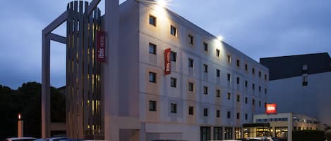 A modern hotel with multiple floors, a parking lot with several cars, and illuminated signage.