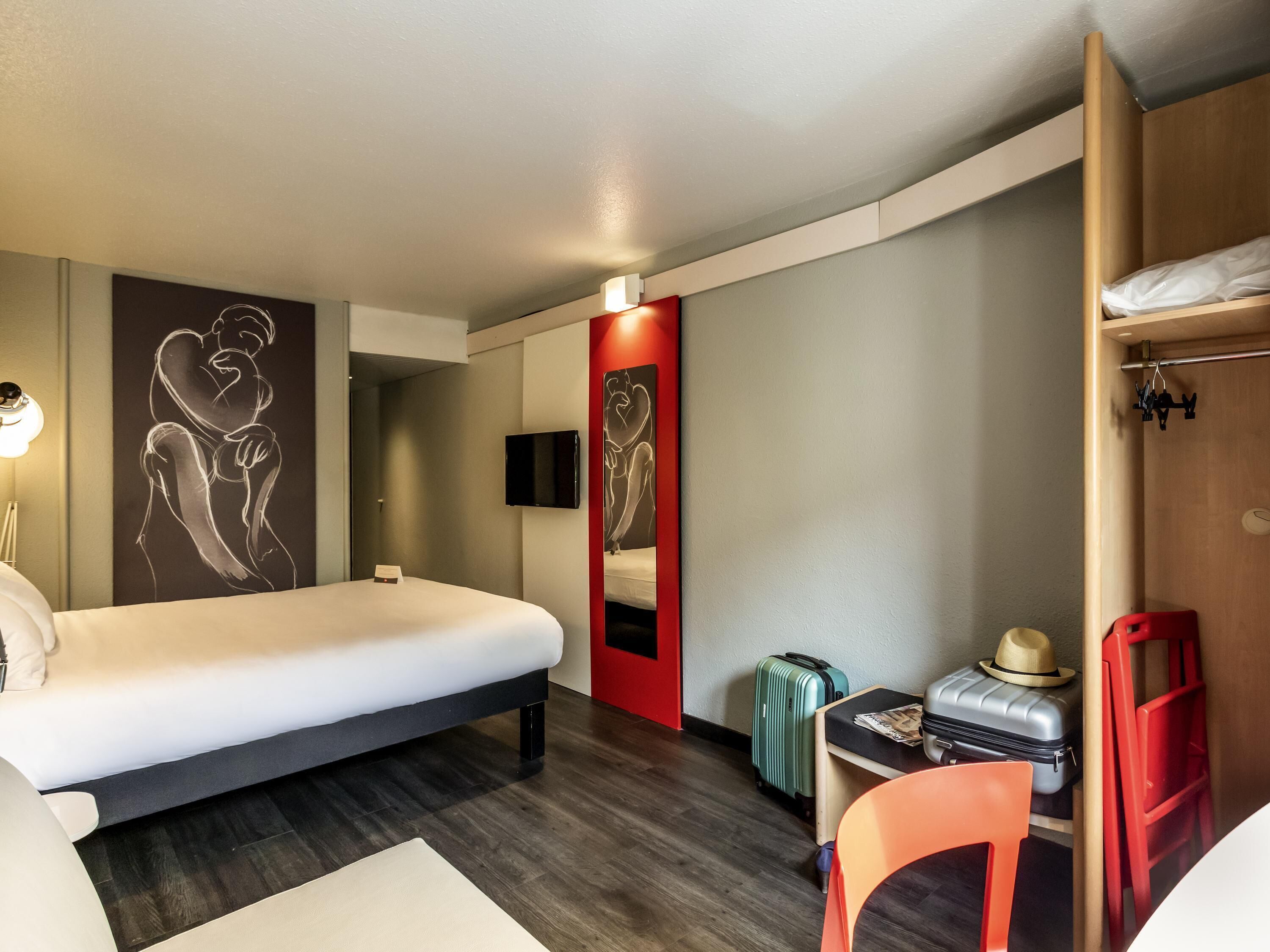 A hotel room with a bed, a desk, a chair, a suitcase, a hat, and artwork on the walls.