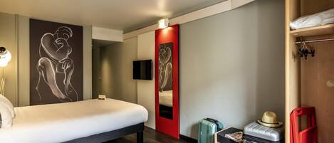 A hotel room with a bed, a desk, a chair, a suitcase, a hat, and artwork on the walls.