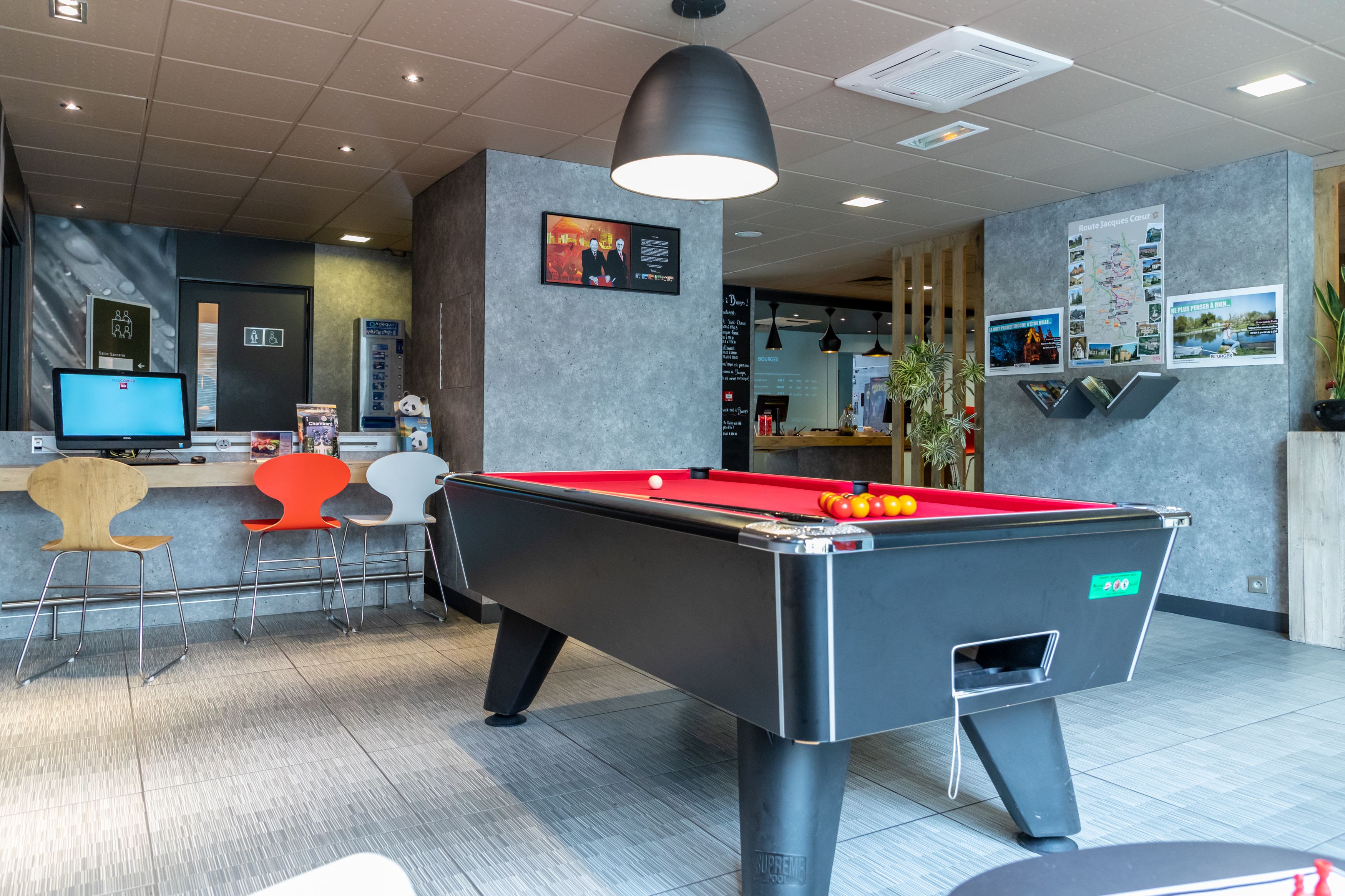 A pool table with a red felt surface, a digital screen, and a seating area with colorful chairs.