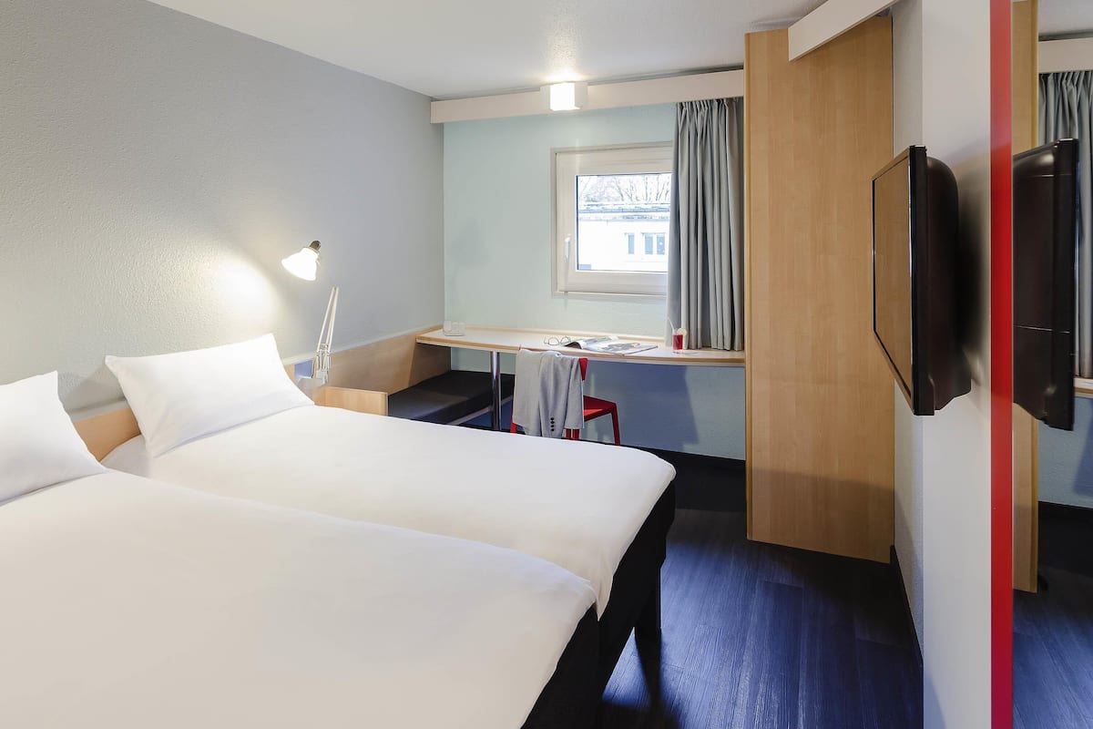 Ein Hotelzimmer mit zwei Betten, einem Schreibtisch, einem Fernseher und einem Fenster mit Vorhängen.