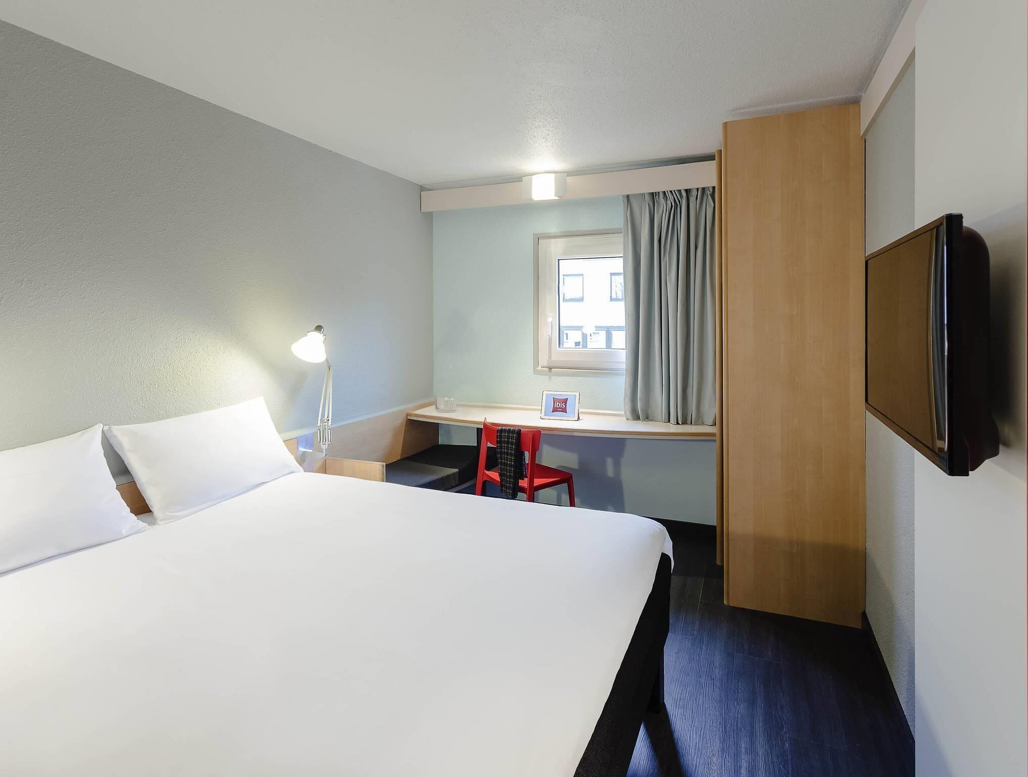 Habitación de hotel con una cama grande, un escritorio con silla, televisión y ventana con cortinas.