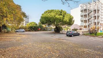 A parking area with two cars, a large tree, and a multi-story building with balconies.