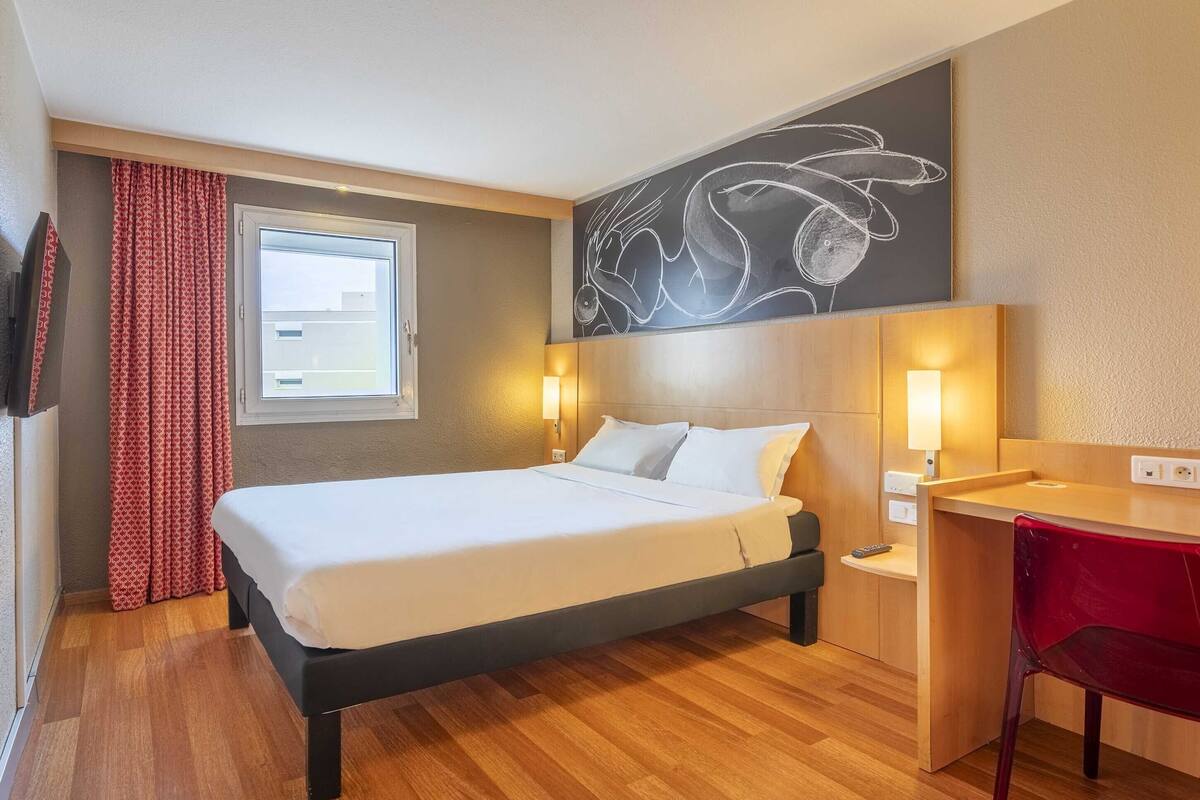 Una habitación de hotel con cama, mesita de noche, lámpara y una silla roja.