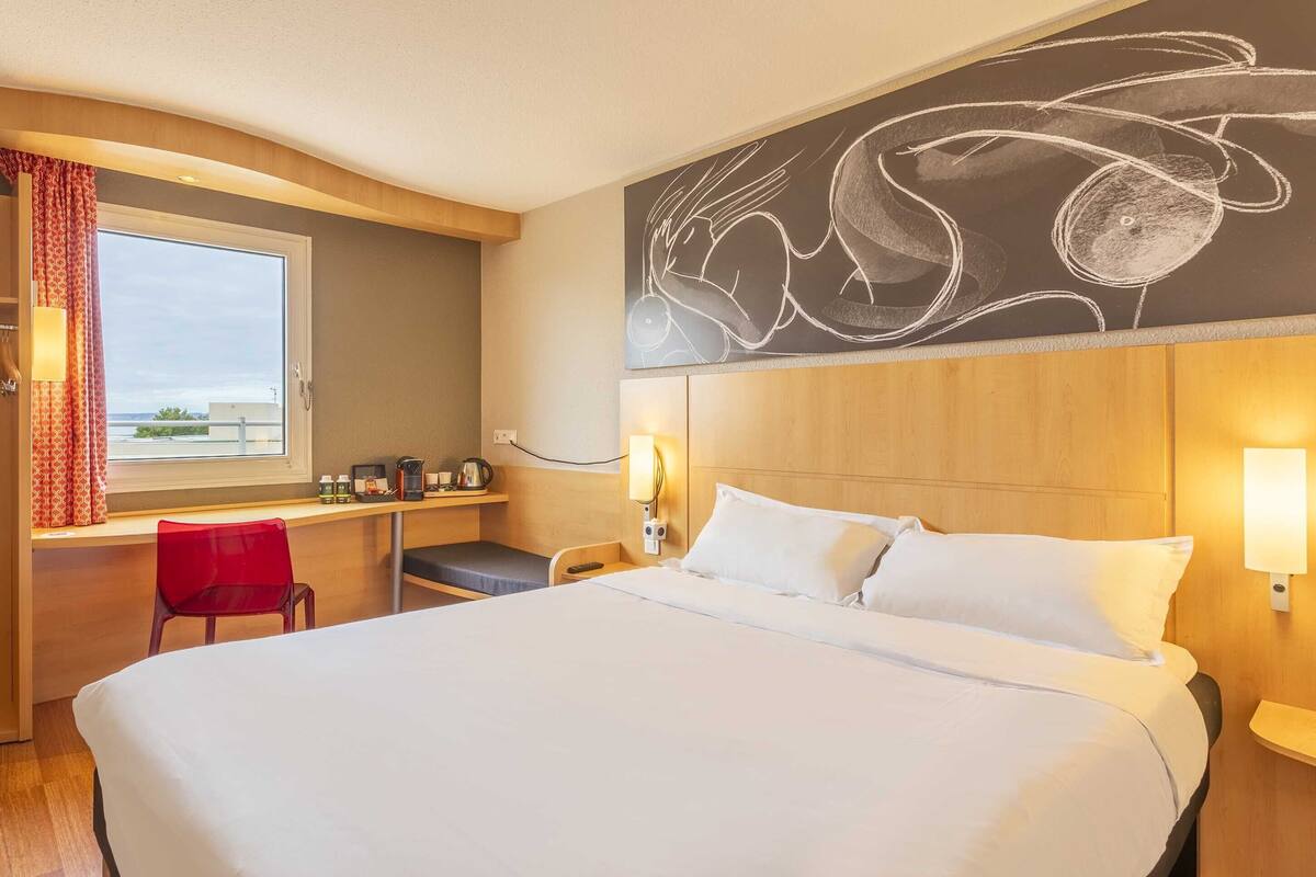 Habitación de hotel con una cama grande, un escritorio con silla, una ventana con cortinas y un pizarrón con un dibujo en la pared.