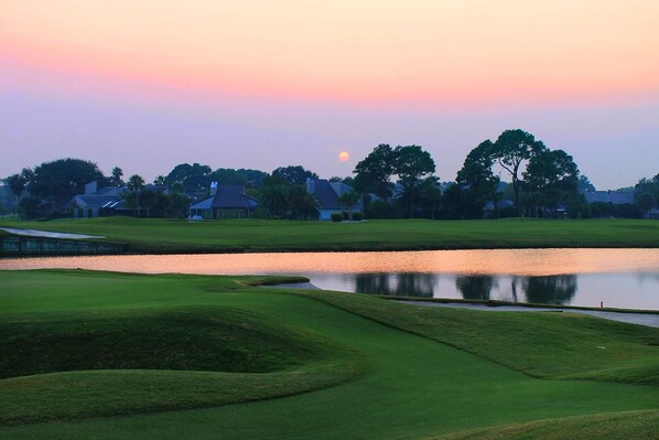 Un campo de golf con un estanque, casas al fondo y un atardecer.