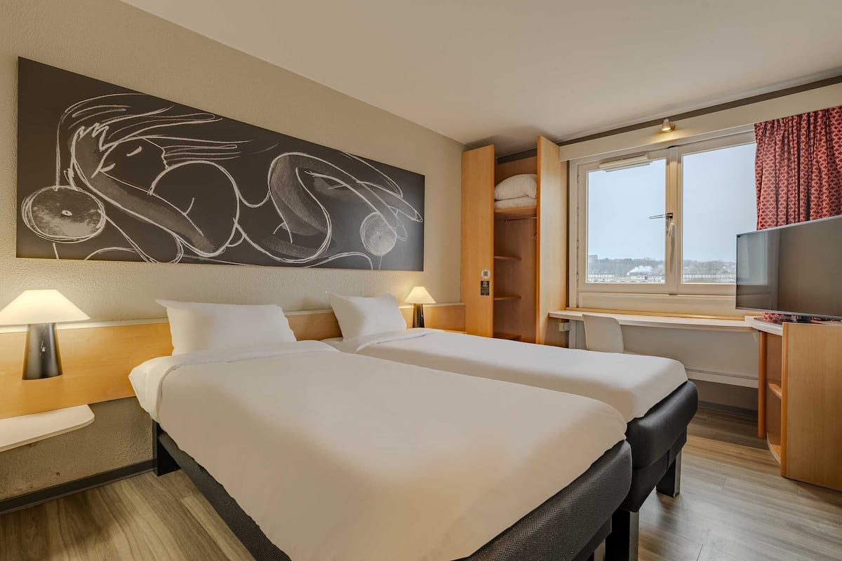 A hotel room with two beds, a large abstract painting, a desk, and a view of the city.