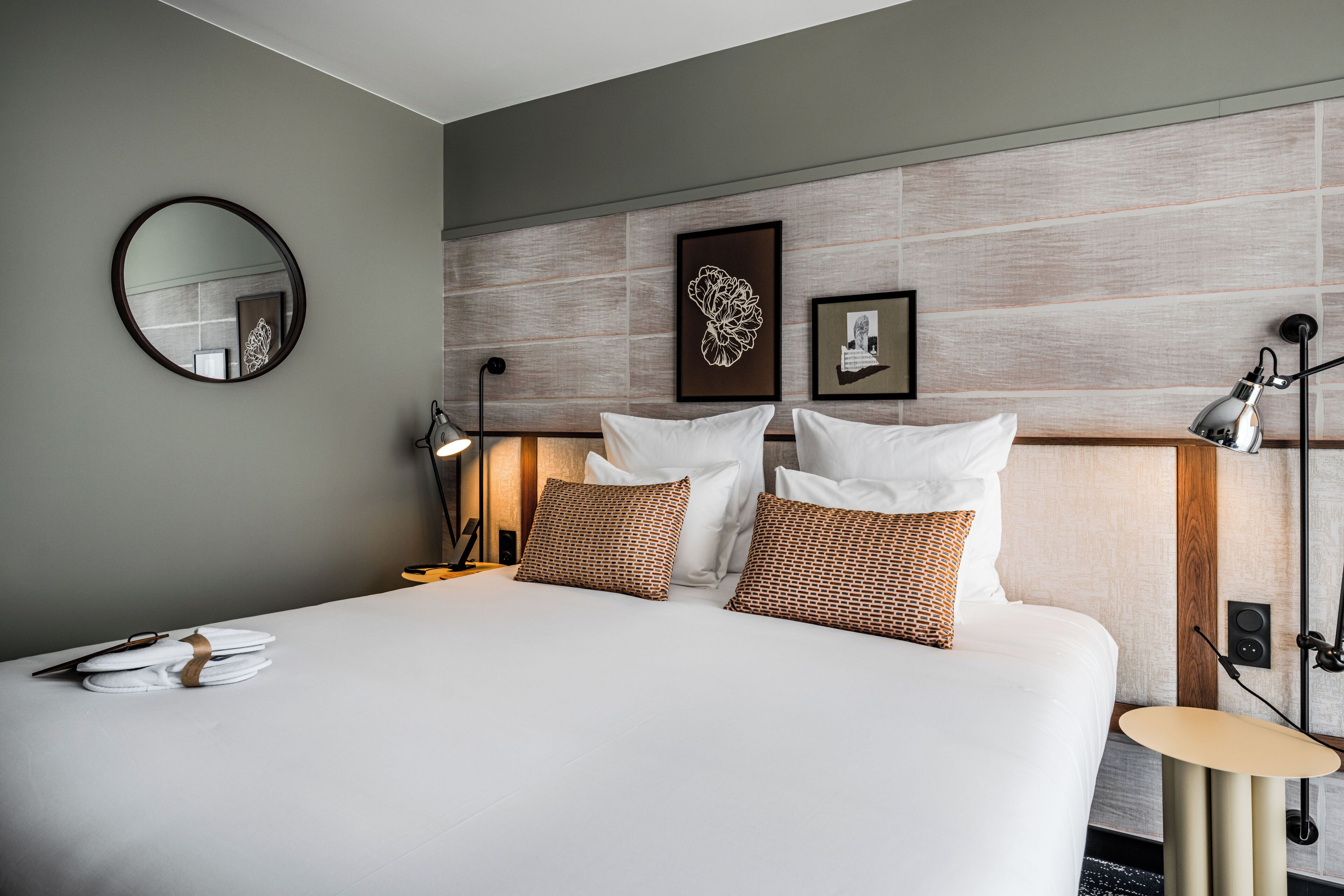 A neatly made bed with white linens and patterned pillows, a round mirror on the wall, and a framed artwork above the bed.