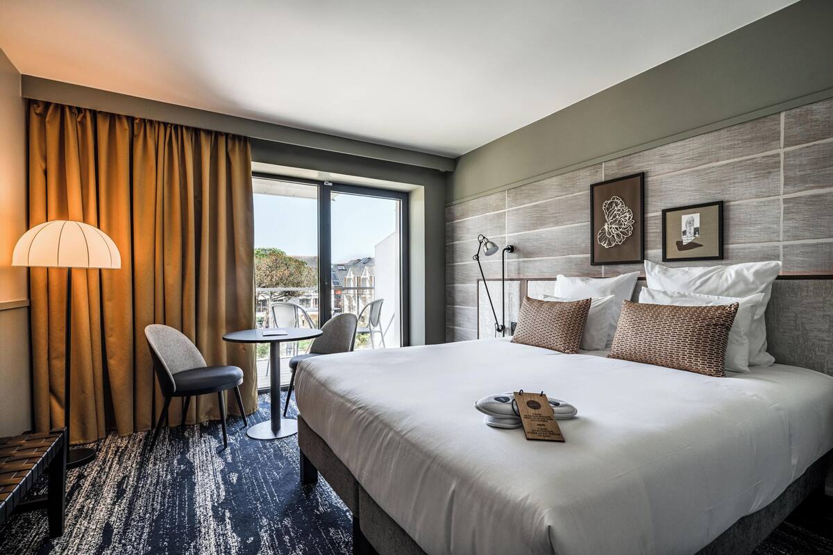 A hotel room with a large bed, a desk with a chair, a lamp, and a view of the outdoors.